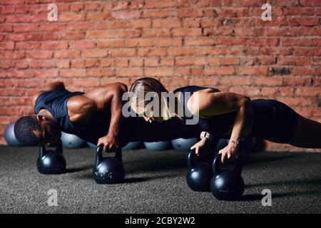 Dunkelhäutiger Mann und weiße Frau, die Push-ups auf Hanteln machen. Cross-fit african Trainer demonstriert Plank Position mit Wasserkocher bellsto seinem Auszubildenden. H Stockfoto