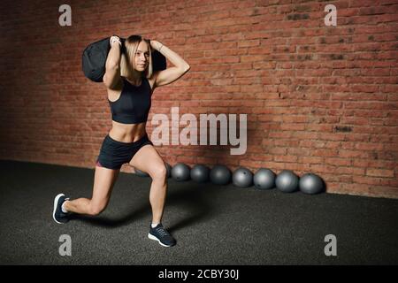 Junge Sportlerin macht Ausfallschritte mit Gewicht, hält in den Händen schweren Sandsack während Schaltung Funktionstraining im Cross-Fit-Studio Stockfoto