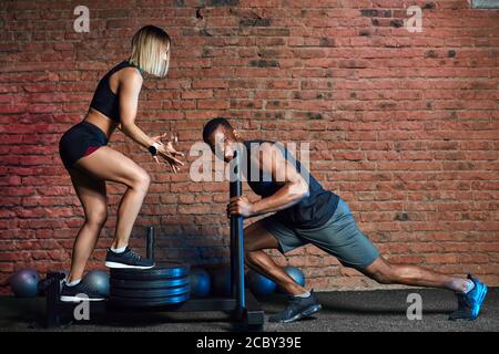 Afrikanischer Athlet, der mit einem schweren Sled Push-Gerät und einer blonden europäischen Frau arbeitet, unterstützt ihn im Indoor-Cross-Fit-Studio. Stockfoto