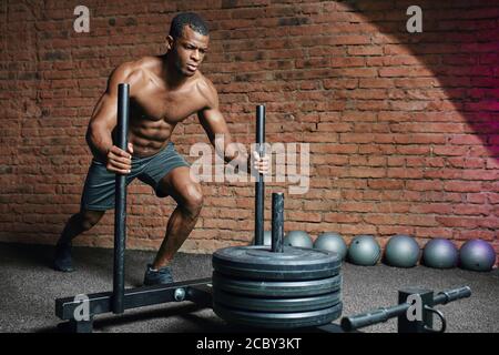 Afrikanischer Sportler schiebt Schlitten in Crosstraining Turnhalle. Muskulösen und starken Mann schieben den Schlitten Cross fit Ausrüstung auf Kunstrasen Rasen. Stockfoto