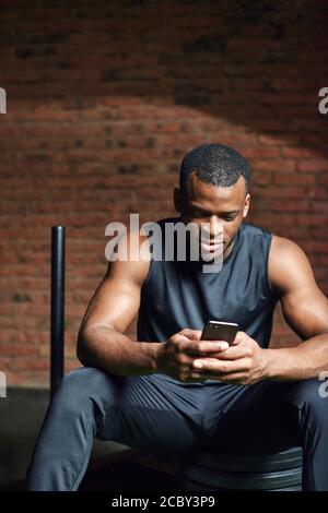 Portrait des afrikanischen Athleten Mann, der sein Smartphone während der Ruhe nach dem Training im Fitnessstudio überprüft. Stockfoto