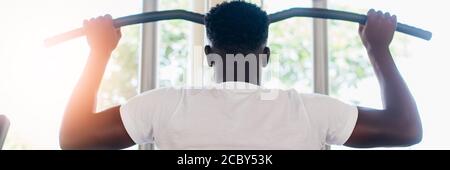 Starker afroamerikanischer Mann in Sportswear, der während des Trainings im Fitnessstudio Kabel mit Bar zieht und auf dem Trainingsgerät sitzt. Stockfoto