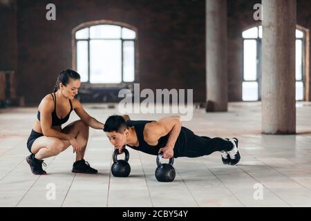 Starker motivierter Mann tut Plank mit Kettlebells während des Trainings im Fitnessstudio, während seine Freundin ihn anschaut. Hobby, Interesse, Motivation Stockfoto