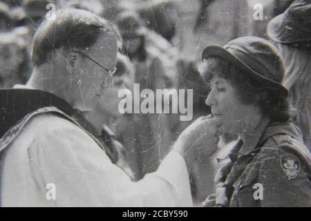 Feine 70er Jahre Vintage Schwarz-Weiß-Fotografie eines Priesters, der die heilige Kommunion bei einer Outdoor-Scouting-Veranstaltung gibt. Stockfoto