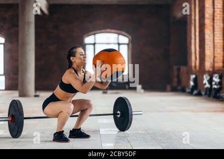 Gut gebaute motivierte Mädchen tun Hocke Übungen mit Fitness-Ball. Langhantel auf dem Boden auf dem Hintergrund des Fotos, Kombination von Kroos fit nackend Stockfoto