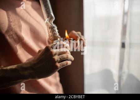 Zugeschnittenes Foto von jungen Mann Heizung Bong mit einem Feuerzeug. Legalisierung von Marihuana, Cannabis, Hanf-Konzept. Mann rauchen zu Hause. Ungesunde Lebensweise, dru Stockfoto