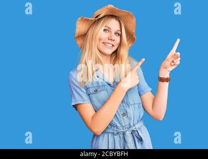 Junge schöne blonde Frau trägt Sommerhut und Kleid lächelnd und Blick auf die Kamera zeigen mit zwei Händen und Fingern zur Seite. Stockfoto