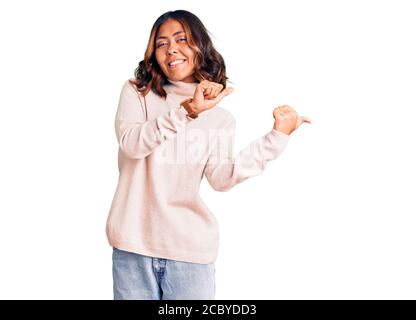 Junge schöne Mixed Race Frau trägt Winter Rollkragenpullover zeigt auf den Rücken hinter mit Hand und Daumen nach oben, lächelnd zuversichtlich Stockfoto