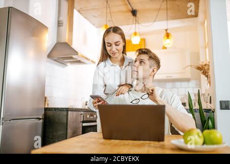 Schöne kaukasische Ehepaar zusammen zu Hause, Mann arbeiten von zu Hause aus, bei Freiberufler. Sorgfältige Frau Unterstützung und umarmen ihn von hinten. Mann verwenden Laptop, sur Stockfoto