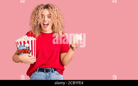 Junge blonde Frau mit lockigem Haar essen Popcorn schreien stolz, feiert Sieg und Erfolg sehr aufgeregt mit erhobenen Armen Stockfoto