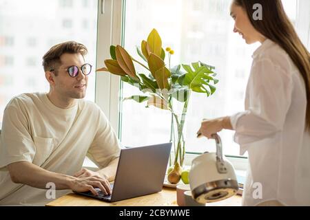 Schöne junge kaukasische Frau Gießen Tee zu arbeiten Ehemann, Mann arbeiten von zu Hause aus mit Laptop. Barmherzige freundliche Frau unterstützen ihn, wollen ihm eine c Stockfoto