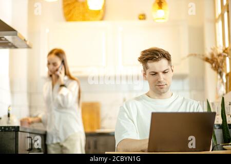 Verheiratet kaukasischen Paar arbeiten von zu Hause aus während Quarantäne, Mann sitzen mit Laptop, während seine Frau löst geschäftliche Probleme per Telefon. Arbeiten online, Freelan Stockfoto