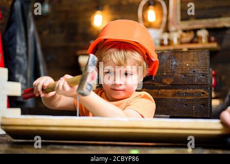 Kid hämmert Nägel mit einem Hammer in ein Holzbrett. Junge Kind beschäftigt im Helm lernen, Hammer zu verwenden. Kind hämmert Nagel in Holz zu Hause. Stockfoto