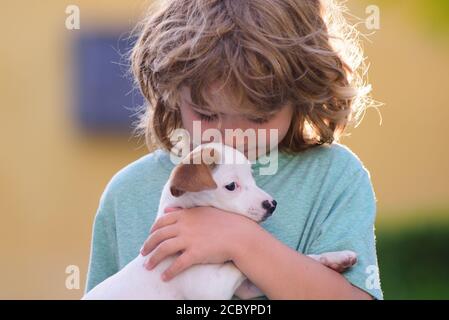 Niedliches Kind Junge fühlt sich erfreut, schließt Augen vor Vergnügen, trägt kleinen Hund, drückt zarte Emotionen, Pflege und Liebe zu kleinen Welpen. Kind mit Stockfoto