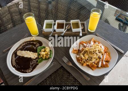 Huevos Oaxaquenos, oder Spiegeleier in schwarzer Maulwurfsauce mit Sesamsamen und geschmolzenem Käse auf einer Maistortilla mit Kartoffeln, links, und Chilaquiles, Stockfoto