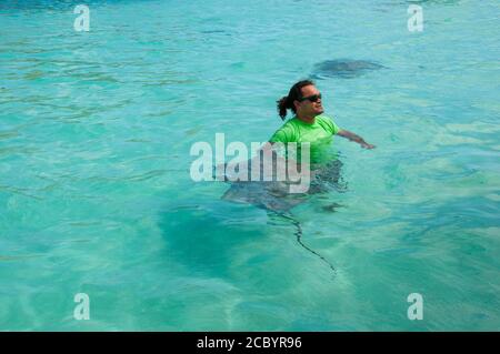 Blick vom Boot aus auf einen polynesischen Mann mit Sonnenbrille, der unanstößige Mantarochen im südpazifik streichelt, während er lächelt und ruhig aussieht. Stockfoto