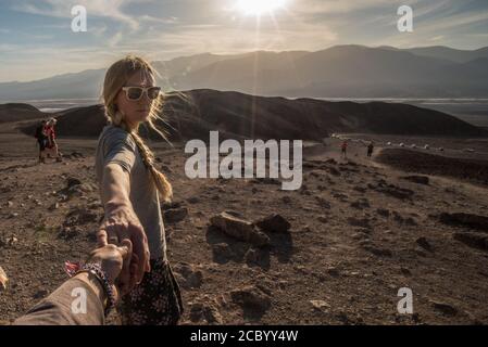 Die blonde Wanderin hält die Hände der Fotografin und blickt zurück in die Kamera, während ihr Haar weht und die Sonne im Death Valley Nationalpark untergeht. Stockfoto