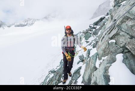 Männlicher Alpinist in Sonnenbrille und Schutzhelm, der festsitzend Seil hält, während er verschneiten Berg klettert. Bergsteiger steigt natürliche Felsformation. Konzept des Winterkletterns. Stockfoto