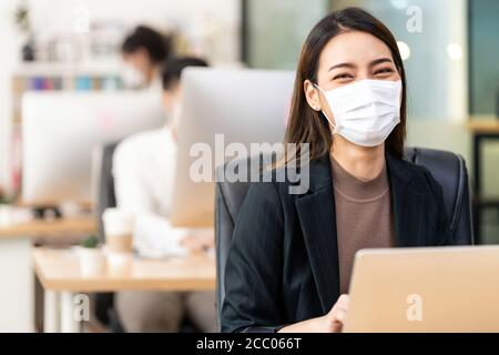 Portrait der asiatischen Büroangestellte Geschäftsfrau tragen Schutzmaske Arbeiten Sie in einem neuen normalen Büro mit interracial Kollegen im Hintergrund Als sozial Stockfoto