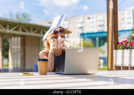 Erwachsene Frau in Brille mit Laptop draußen in der Stadt, versteckt vor der Sonne. Müde Senior suchen Abstand, arbeiten und trinken Kaffee. Fernunterricht Stockfoto