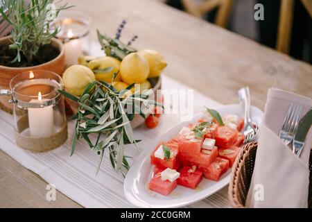 Ein Teller mit Wassermelone in Quadrate geschnitten mit einer tiefen Schüssel mit Zitronen auf dem Tisch mit einer brennenden Kerze. Stockfoto