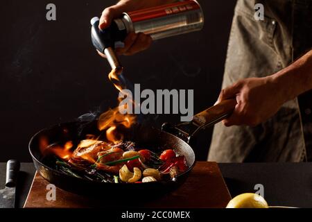 Koch kocht mit Feuer in einer Pfanne Garnelen mit Gemüse. Auf schwarzem Hintergrund Stockfoto