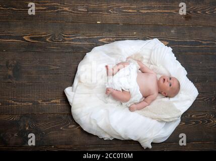 Kindheit und Göttliche Geschenk Konzept. Kleinkind mit weißer Bettdecke bedeckt. Neugeborenes Kleinkind mit blauen Augen und neugierigem Gesicht auf Holzhintergrund. Baby liegt auf weißen weichen Decke als Wolke oder Nest entworfen Stockfoto