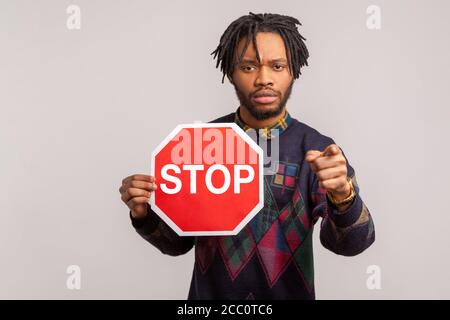 Halt! Besorgt besorgt afrikanischen Kerl mit Dreadlocks halten Stop-Schild und zeigen Finger auf die Kamera mit ernst ängstlich Gesicht, aufgeben betrunken fahren. Stockfoto