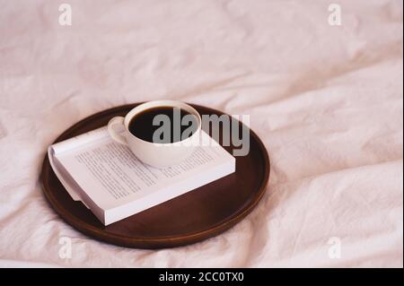 Tasse frischen Kaffee auf offenem gefaltetem Buch auf Holztablett im Bett in Nahaufnahme. Guten Morgen. Stockfoto