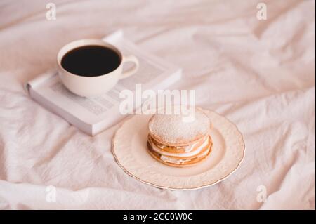 Tasse frischen Kaffee auf offenem Buch mit Pfannkuchen auf weißem Teller im Bett Nahaufnahme. Guten Morgen. Selektiver Fokus. Stockfoto