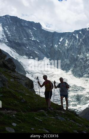 Zinal, Schweiz - 19. Juli 2019: Volle Länge der Reisenden mit Trekkingstöcken zu Fuß entlang Hügel grasbewachsenen Weg mit schneebedeckten Kamm im Hintergrund. Alte Dame und Herr schlendern die Bergstraße hinunter Stockfoto