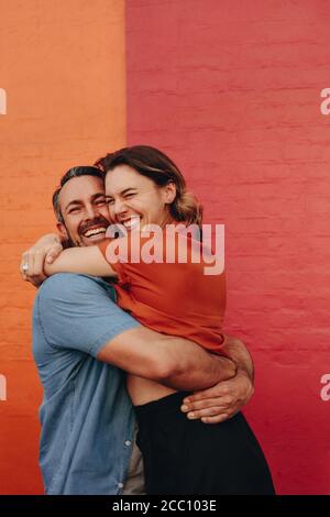 Liebevolles Paar mittleren Alters umarmt gegen farbige Wand. Reifer Mann und Frau zusammen gegen rote und orange Wand. Stockfoto