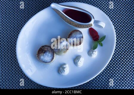 Drei Hüttenkäse Pfannkuchen bestreut mit Puderzucker auf einem weißen Teller einer Führungsform. Kleines Glas Marmelade Stockfoto