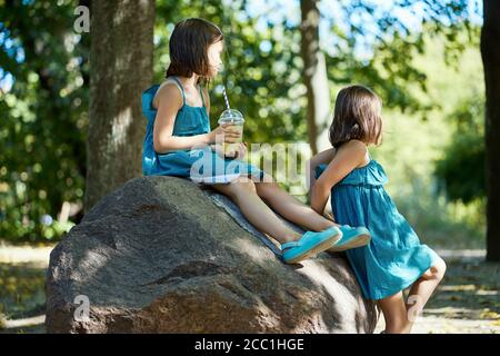 Zwei niedliche kleine Mädchen in Kleidern sitzen auf einem Stein Und Limonade trinken Stockfoto