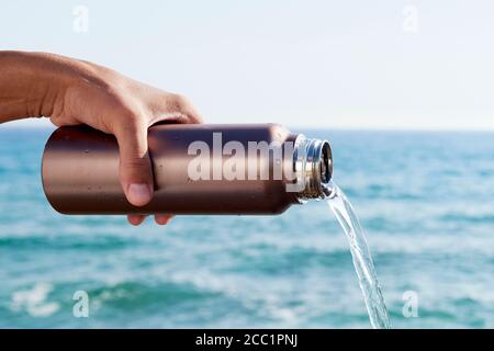 Nahaufnahme eines Mannes, der Wasser aus einem wiederverwendbaren Aluminium gießt Wasserflasche vor dem Meer Stockfoto