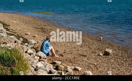 Frau saß allein, mit Blick auf das Meer, auf einem Kiesstrand am Hafen von Emsworth, Hampshire, Großbritannien. Stockfoto