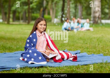 In voller Länge Porträt von niedlichen Mädchen von amerikanischen Flagge bedeckt sitzen auf Picknick-Decke im Park und lächeln an der Kamera, kopieren Raum Stockfoto