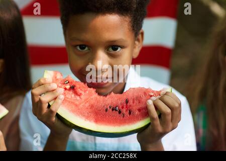 Nahaufnahme Porträt eines lächelnden afrikanischen Jungen hält Wassermelone und Blick auf die Kamera mit amerikanischer Flagge im Hintergrund, kopieren Raum Stockfoto