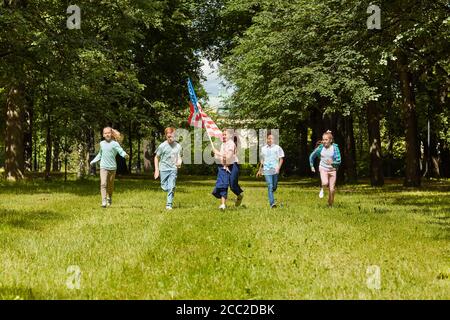 Weitwinkel-Ansicht auf multi-ethnische Gruppe von Kindern, die auf Kamera über grünen Rasen mit Mädchen winken amerikanische Flagge, Kopierer Raum laufen Stockfoto