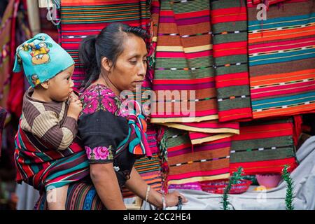 Einheimische Maya K'iche Frau mit Kind auf dem Rücken verkauft Stoff am Markttag in der Stadt Chichicastenango, El Quiché, Guatemala, Mittelamerika Stockfoto