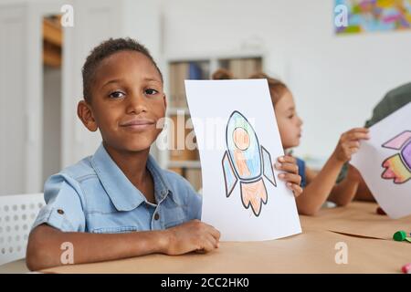 Portrait von niedlichen afroamerikanischen Jungen zeigt Bilder von Weltraumrakete Und lächelnd auf die Kamera, während Sie Kunstunterricht in Pre genießen Schule oder Kinder develo Stockfoto