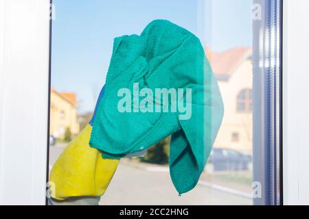 Eine Hand in einem gelben Handschuh wischt das Fenster mit einem Lappen vor dem Hintergrund von niedrigen Gebäuden. Stockfoto