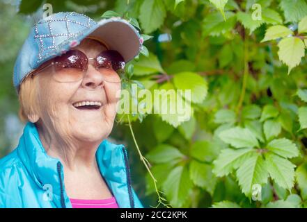 Fröhliche, modische 90-jährige Großmutter mit grauen Haaren, Sonnenbrille und einem Lächeln in der Natur an einem sonnigen Sommertag. Stockfoto