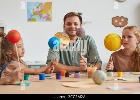Porträt eines lächelnden männlichen Lehrers, der Planetenmodelle hält und die Kamera anschaut, während er mit einer Gruppe von Kindern in Kunst- und Bastelunterricht arbeitet, Raum kopieren Stockfoto