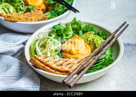 Buddha-Schüssel mit Zucchini-Pasta, gegrilltem Tofu, Guacamole, Süßkartoffel-Hummus und Gemüse, grauer Hintergrund. Stockfoto