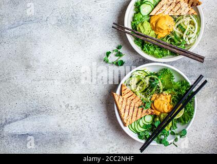 Buddha-Schüssel mit Zucchini-Pasta, gegrilltem Tofu, Guacamole, Süßkartoffel-Hummus und Gemüse, grauer Hintergrund. Stockfoto