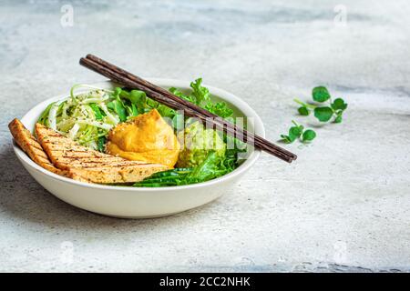 Buddha-Schüssel mit Zucchini-Pasta, gegrilltem Tofu, Guacamole, Süßkartoffel-Hummus und Gemüse, grauer Hintergrund. Stockfoto