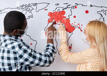 Rückansicht von zwei verschiedenen Menschen, die auf neue Infektionsfälle in der Welt hinweisen, rote Krankheitszeichen auf der Weltkarte. Coronavirus, COVID-19 Konzept Stockfoto