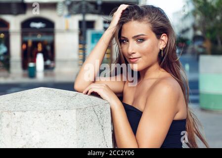 Junge Frau mit hellen Augen und blonden Haaren in einem städtischen Gebiet, auf einer Seite des Fotos auf Betonblock gelehnt, mit einer Hand auf dem Kopf lächelnd Stockfoto