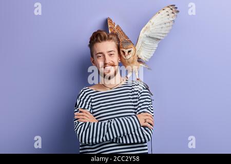 Fröhlicher positiver junger Mann mit einer Eule in der Schulter, die fliegen wird. Isolierter blauer Hintergrund, Studioaufnahme. Stockfoto